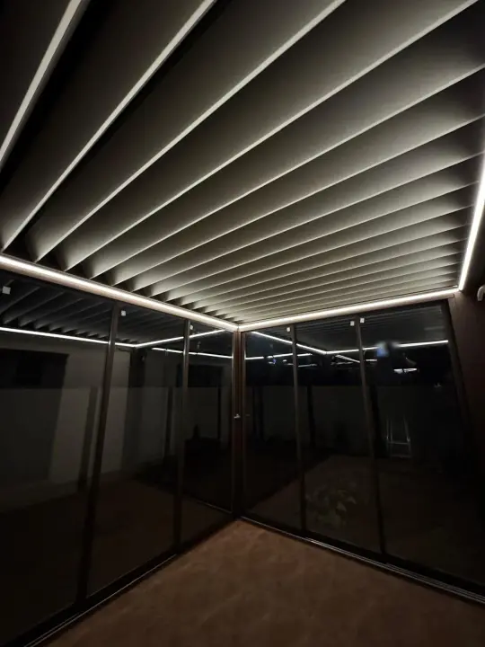 Corner of the glass-enclosed terrace at night, adjustable louvres open and perimeter LED on